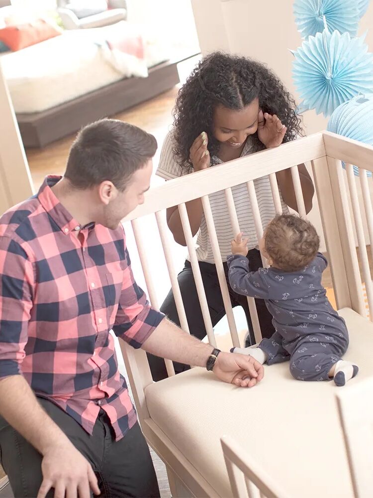 A woman and man play peek-a-boo with a baby in a crib