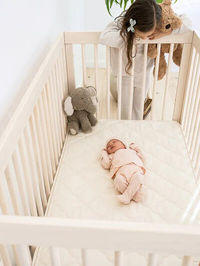 A baby lies on a mattress while a child looks at it from over the top of the crib.