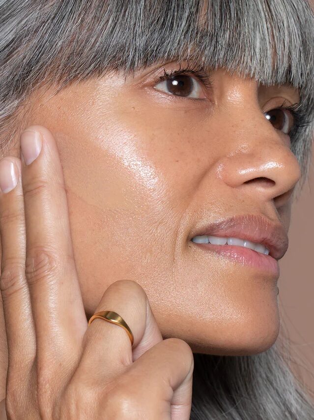 A close up of a model with gray bangs shows her swiping the product on her cheek bone.