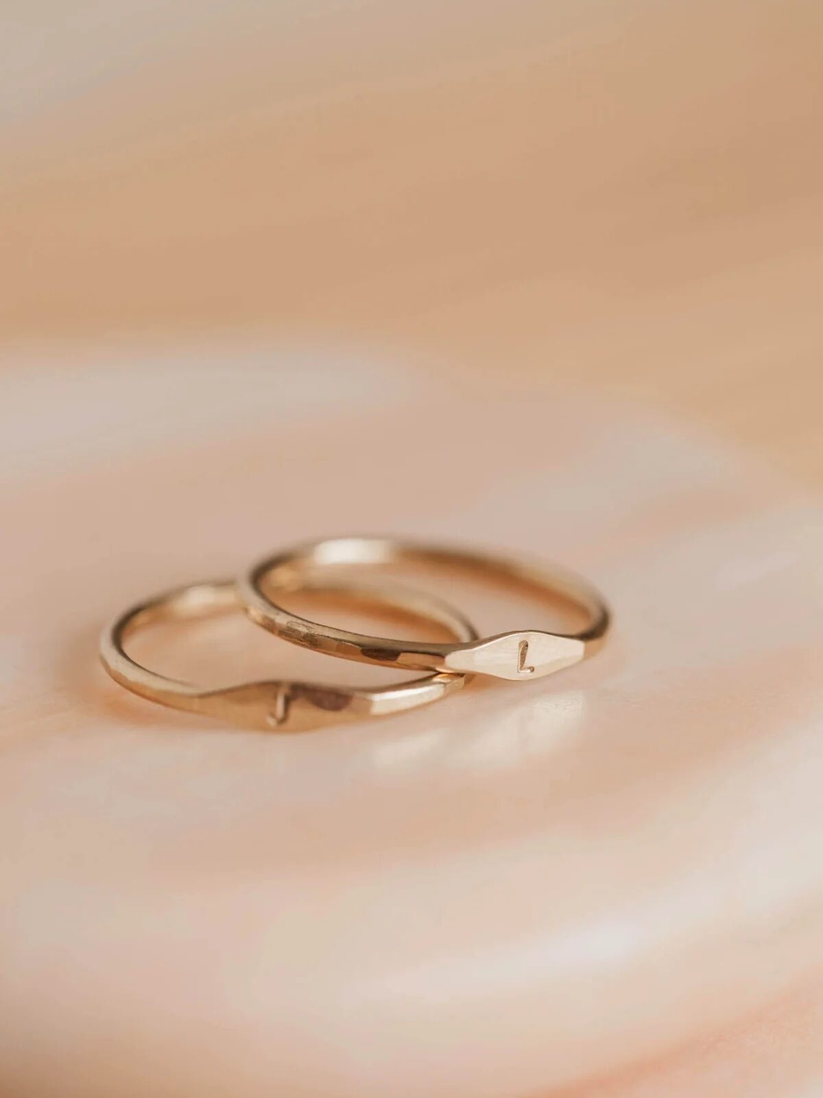 Two small gold signet rings with the letters "L" and "J" are stacked on a table.