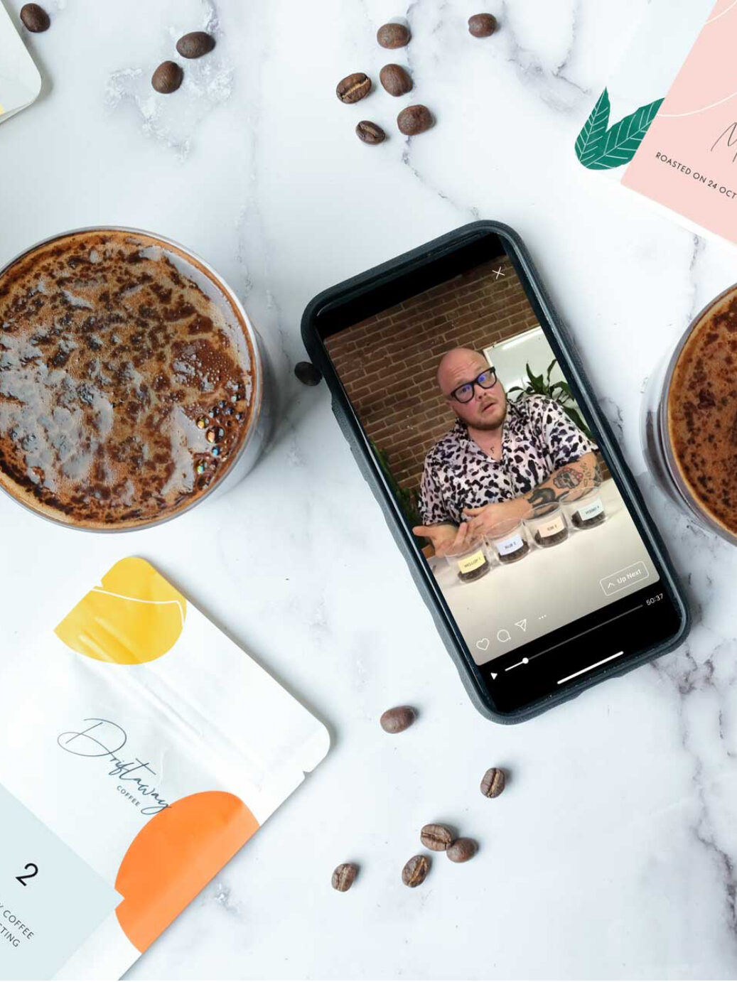 A marble top table with loose coffee beans, a brewing cup of coffee, a Driftaway coffee bag, and a virtual tasting on a phone screen.
